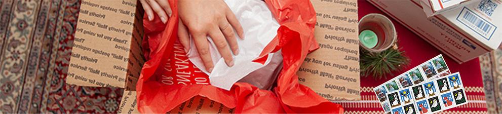 一个人正在包装一个假日包裹，桌上放着优先邮箱和假日邮票.